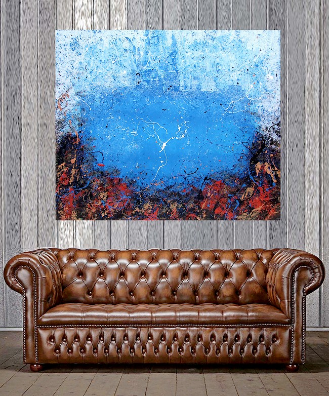 UN ELAGANTE DIVANO CHESTERFIELD DI LUSSO IN PELLE MARRON CHIARO CON BORCHIE SOPRA AD UN PAVIMENTO IN TAVOLE DI LEGNO DOGATE E SULLA PARETE METALLICA APPESO UN QUADRO DI GOA COLOR CELESTE, AZZURRO, ROSSO E BIANCO