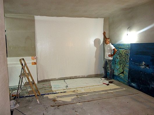 L'ARTISTA NEL STUDIO CON LA GRANDE TELA APPESA AL MURO